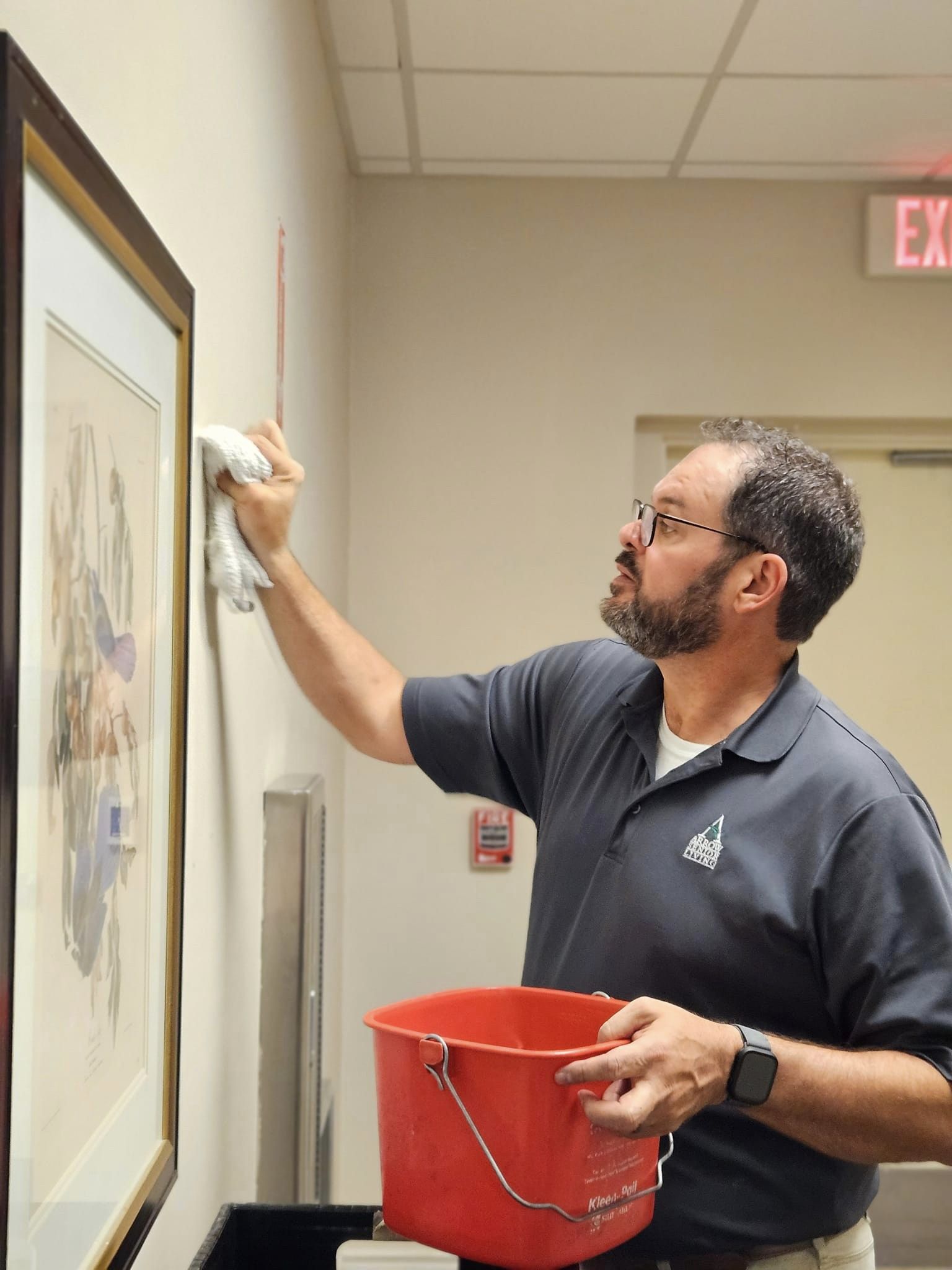 A male staff member wearing a gray polo shirt is carefully cleaning a framed picture on a wall, holding a red bucket in one hand, in a well-lit hallway with an exit sign.