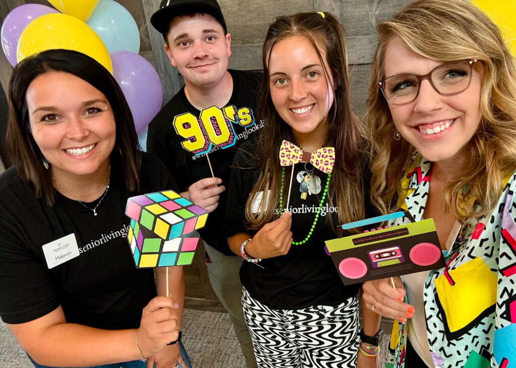 Four smiling people dressed in 90s-themed attire, holding fun props like a Rubik's cube, a '90s' sign, and a boombox cutout, posing for a photo at a job fair event.