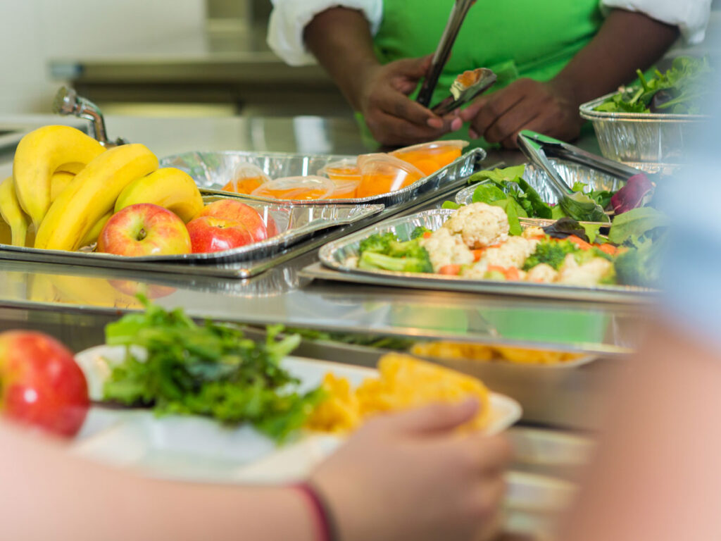 School cafeteria food line with trays of fresh fruits, salads, and vegetables being served by a cafeteria worker in a green apron.