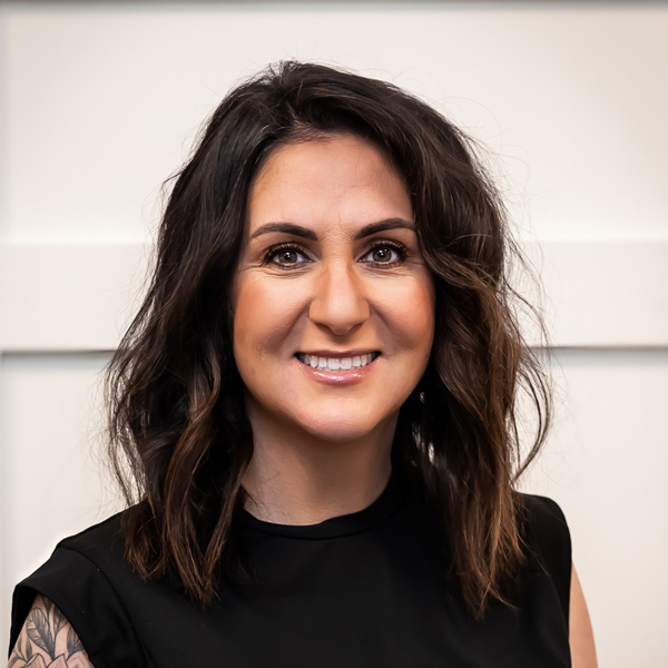 Allie Ebersole, Business Office Director at PrairieStone Senior Living, smiling in a black sleeveless top, with wavy dark hair, standing against a white paneled background.