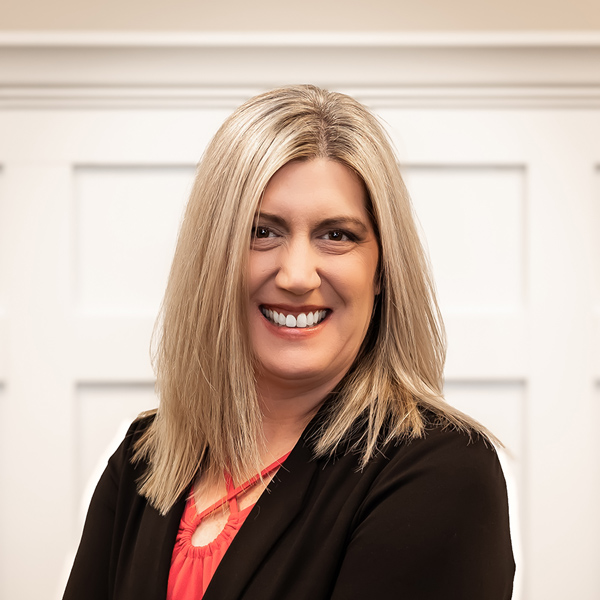 Michelle 'Micki' Brodhead, Operations Coordinator at PrairieStone Senior Living, smiling warmly in a black blazer and red blouse, standing against a white paneled background.
