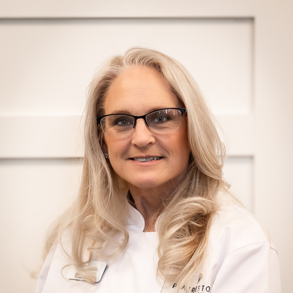 Robyn Beenken, Culinary Director at PrairieStone Senior Living, wearing a white chef's coat and glasses, with long wavy blonde hair, smiling against a white paneled background.