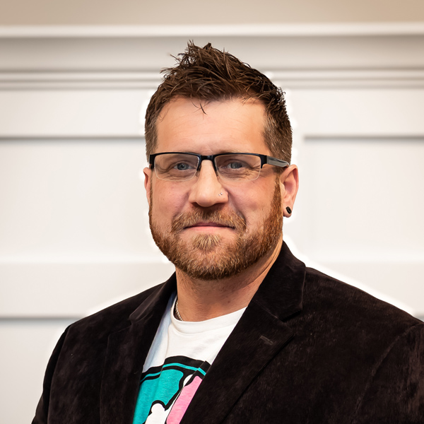 Todd Sherrets, Plant Operations Director at PrairieStone Senior Living, wearing glasses, a black blazer over a graphic T-shirt, and sporting a short beard, standing in front of a white paneled wall.